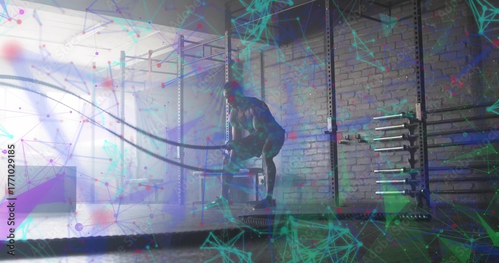 Fototapeta premium Swinging athlete performing battle rope exercise in gym, with metal rig, barbells and weight plates