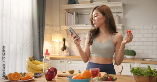 Young Asian woman in sportswear enjoys organic healthy fruits in kitchen while checking health data on smartphone, using AI-powered nutrition or wellness app to support clean diet balanced lifestyle.