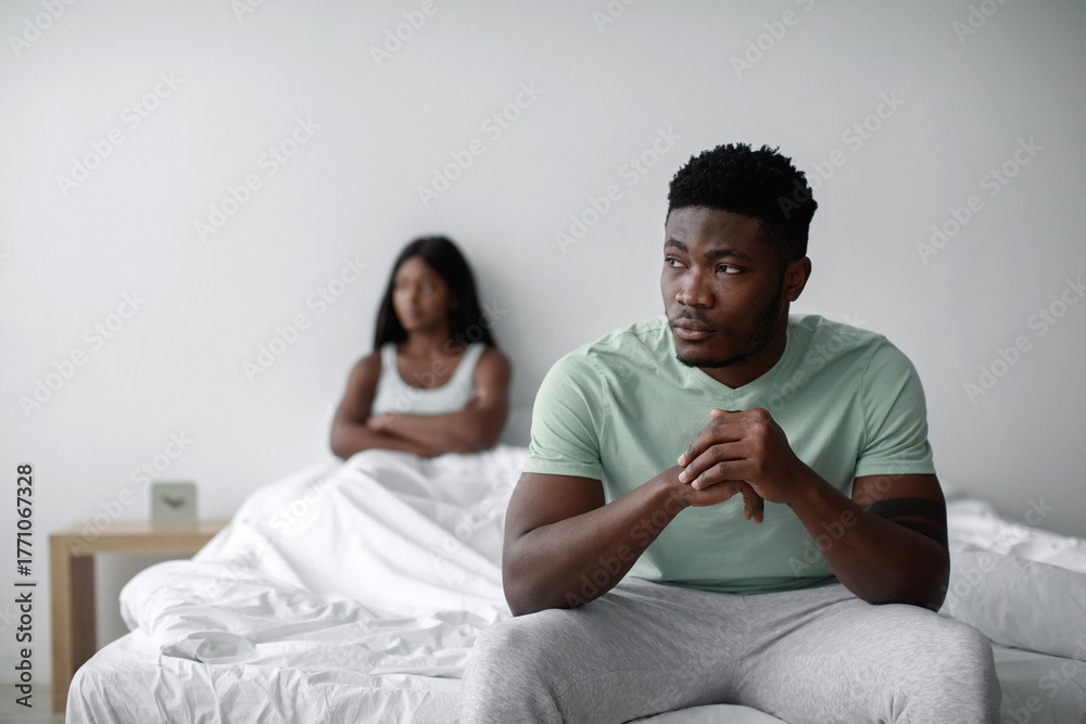 Obraz premium A man sits on the edge of a bed looking pensive while a woman in the background sits with her arms crossed, indicating emotional distance in a quiet setting.