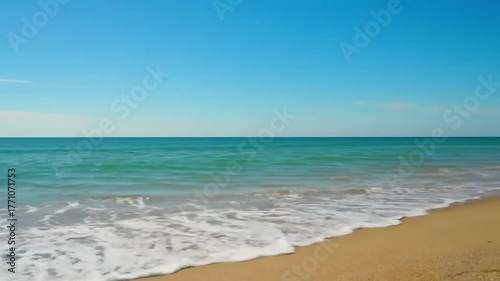 A serene beach scene with calm, turquoise waves gently lapping the shore, golden sand stretching out under a clear blue sky, with a distant view of the ocean horizon, medium shot, slow camera pan.
