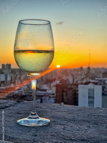 A glass of white wine rests on a stone railing overlooking a breathtaking view of Buenos Aires at sunset. The golden light illuminates the cityscape, creating a warm and elegant atmosphere.