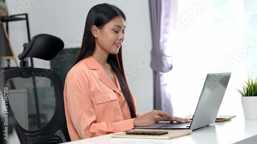 Asian young woman working laptop home desk wearing peach blouse with notebook and smartphone nearby smiling while typing bright modern office ergonomic chair beside potted plant natural light focused