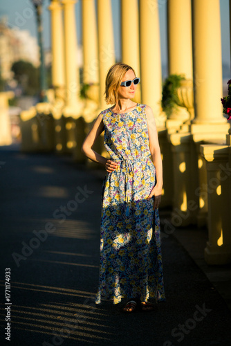 A woman in a summer dress and sunglasses walks along the seaside promenade by the ocean. The bright scene captures lightness, freedom, and the warmth of a coastal day.