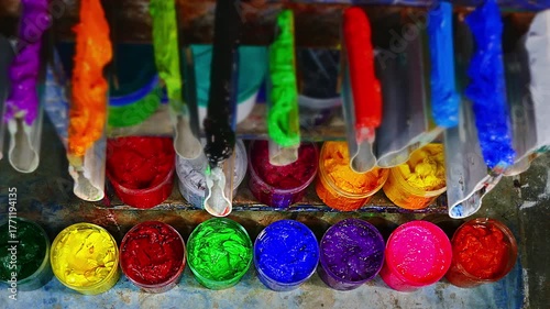 On a wooden workbench in a screen printing shop, several plastisol ink cans are open, revealing rich tones soft pastels, strong primaries, and deep shades. A wooden spoon and trowel rest nearby.