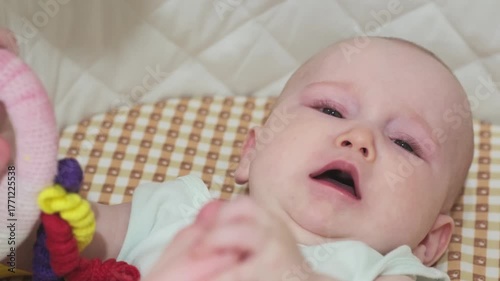  baby girl is lying on the bed and crying. The kid holds her toy and touches her leg.