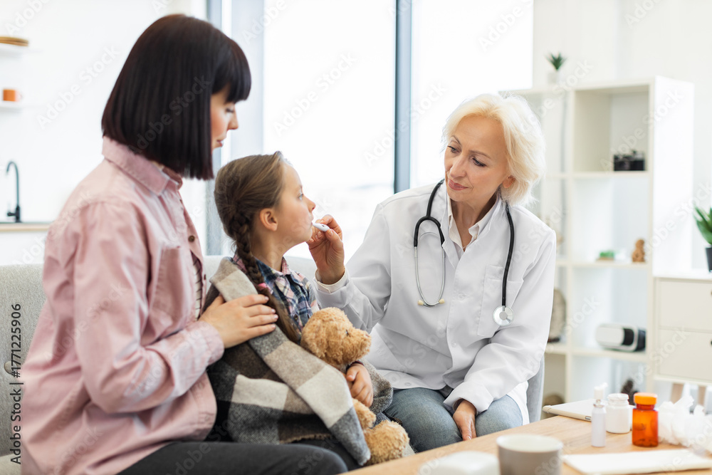 Obraz premium A female doctor checks a child's temperature while the mother comforts her in a doctor's office setting. The girl is wrapped in a blanket with a teddy bear.