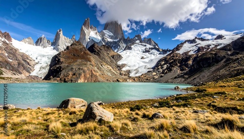 Lake and craggy mountains with glacial caps under a partly cloudy sky, on a sunny day with grassy foreground