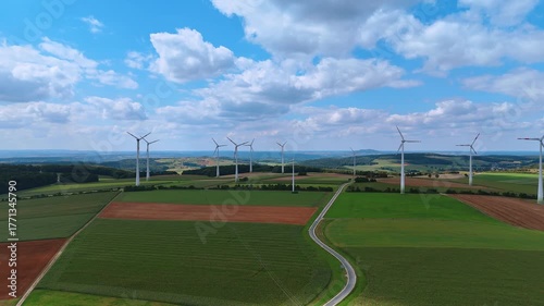 Wind farm over cultivated fields with cloudy sky. Drone landscape view of multiple wind turbines on green fields and brown soil in summer countryside.