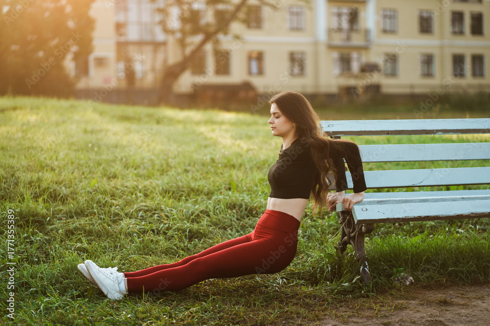 Fototapeta premium A young woman exercises on a park bench under the warm glow of the setting sun, combining fitness with relaxation in a serene urban park setting.