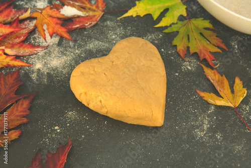 Heart-shaped shortcrust pastry on a floured dark surface, surrounded by autumn maple leaves — cozy seasonal baking and fall-inspired homemade cookie preparation moment.
