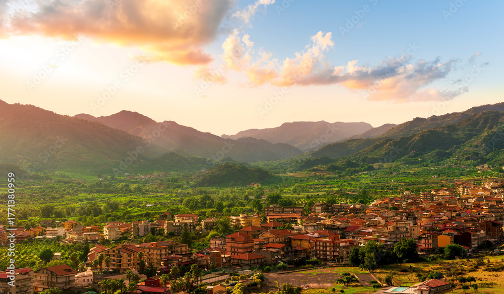Obraz premium wonderful landscape of sunset valley town with beautiful fields, gardens, green plantations among great mountains and scenic evening cloudy sky on background of landscape
