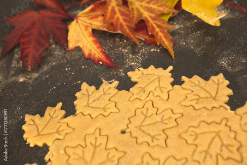 Shortcrust pastry being cut into autumn leaf shapes on a floured dark surface, surrounded by colorful fall leaves — cozy seasonal cookie preparation and festive baking process.
