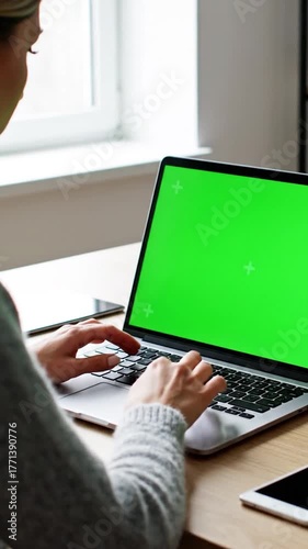 Woman types on laptop with green screen, hands close up, near a window, plus signs on the screen