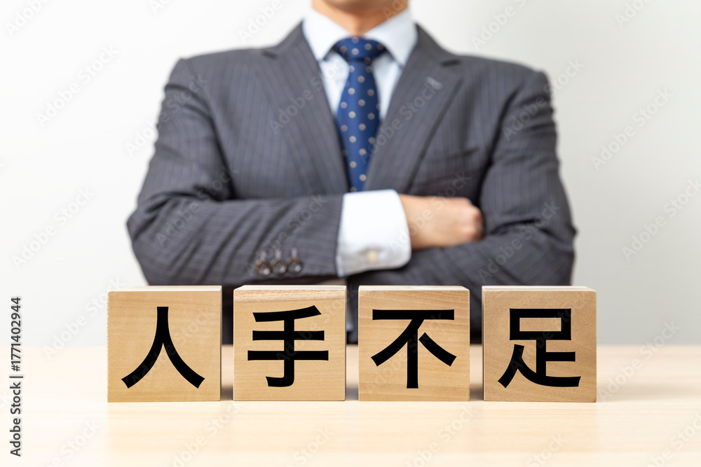 Obraz premium 「人手不足」と書かれたブロックと腕組みをするビジネスマン