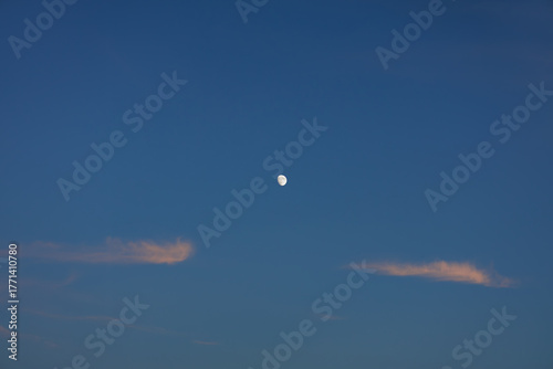 Serene moon hangs high in clear dusk sky, flanked by soft wispy clouds showing hint of pink hue from sunset's scattering sunlight.