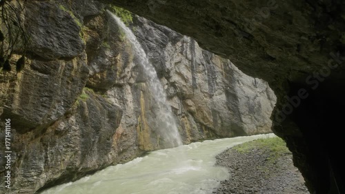 Waterfall cascading into a gorge river in the Swiss Alps, viewed from inside a cave. Dramatic mountain landscape and scenic nature scene