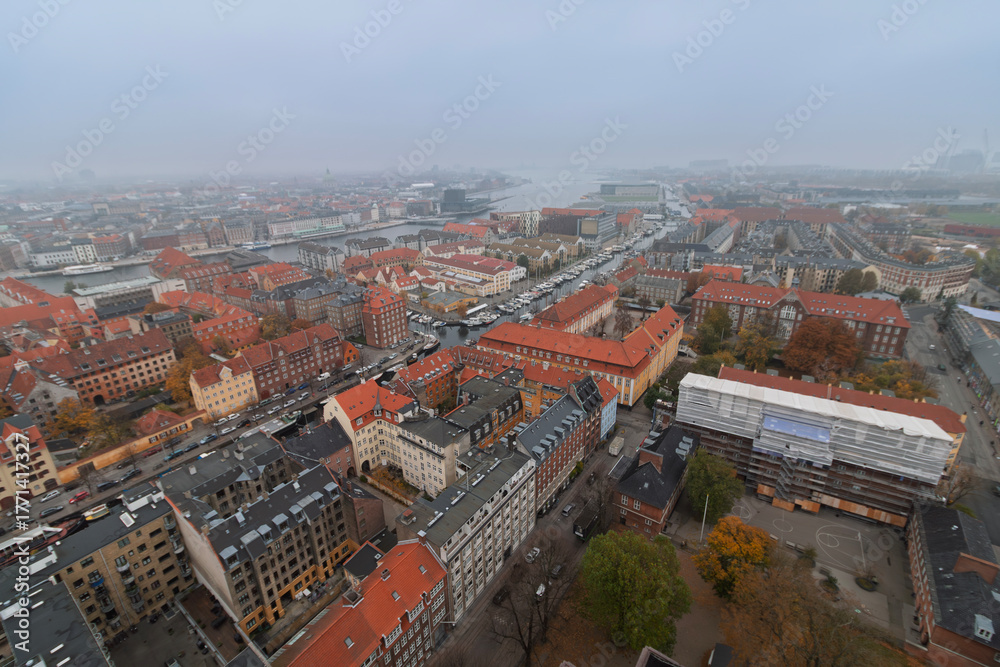 Naklejka premium Copenhagen - An aerial view reveals a dense urban landscape with historic buildings, a waterway lined with boats, and a hazy sky.