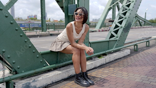 Pretty Asian woman sits on a windy day on a metal bridge, she wears a white sleeveless top paired with a beige skirt and black combat boots