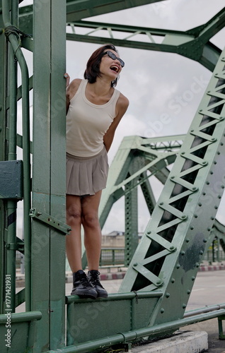 Cheerful Asian woman wearing a beige sleeveless tank top, a short pleated khaki skirt and black combat boots poses playfully on a green metal bridge, exuding a trendy and modern vibe