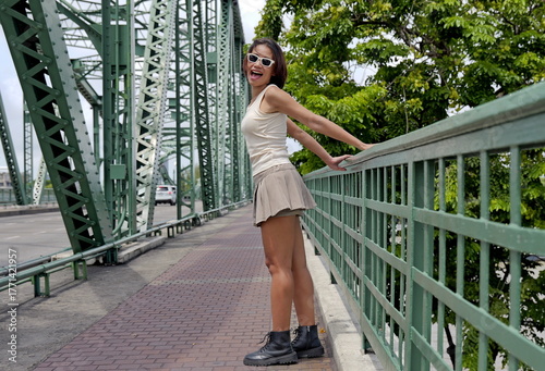 Joyful Asian woman in a beige tank top, pleated skirt and black combat boots stands on a green bridge, showcasing urban fashion and confidence