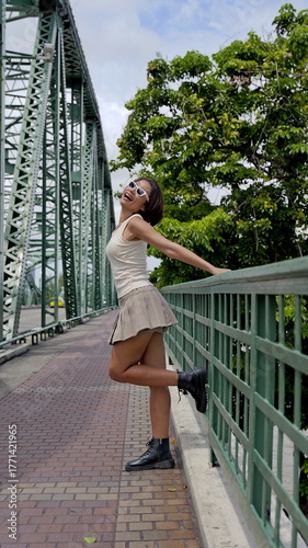 Playful Asian woman in a beige tank top, pleated skirt and black combat boots stands on a green bridge, showcasing urban fashion and confidence
