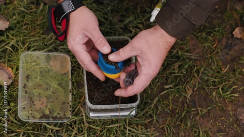 Fisherman preparing fishing feeder with bait, close-up of hands filling feeder mold with groundbait mixture outdoors, detailed angling process and professional fishing equipment concept