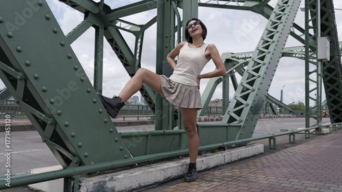 Stylish young Asian woman poses dynamically on a green metal bridge with one foot resting on a beam she is wearing a beige sleeveless tank top, a short pleated khaki skirt, and black combat boots