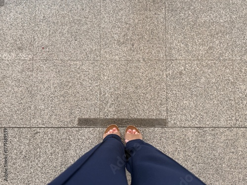 A high-angle,shot of a person's feet wearing brown leather sandals on a light gray,tiled granite or concrete sidewalk with a dark line marking a seam or gutter running across the middle.