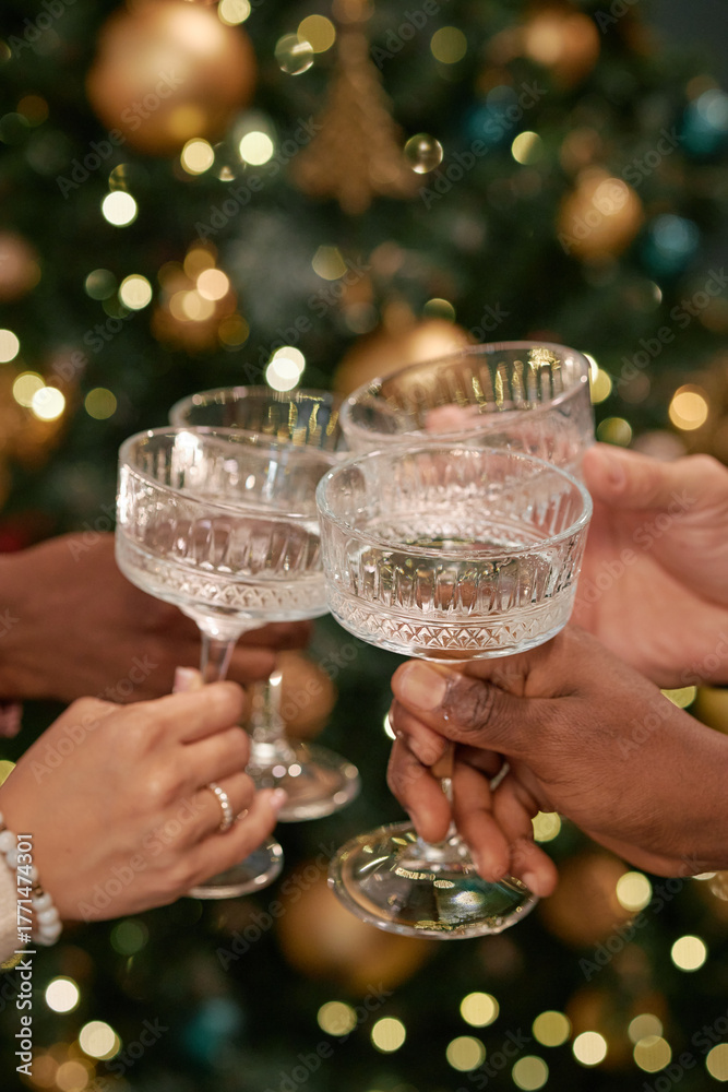 Fototapeta premium Diverse group of young adult men and women clinking crystal glasses in front of decorated Christmas tree, celebrating festive holiday together, hands visible only