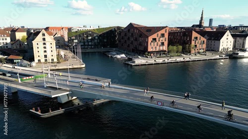 Aerial view of a modern bridge spanning dark waters, connecting red brick buildings under a bright sky, a blend of urban design, Copenhagen, Denmark.