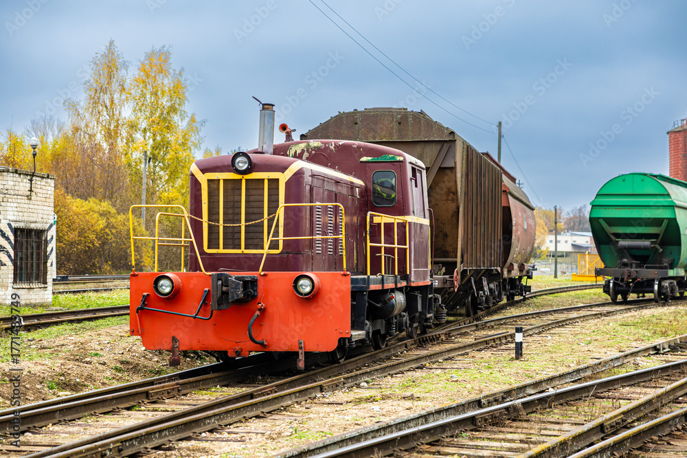 Fototapeta premium A maroon and red diesel locomotive hauls rusty and green freight wagons through a rail yard on an overcast autumn day, surrounded by yellow foliage and tracks