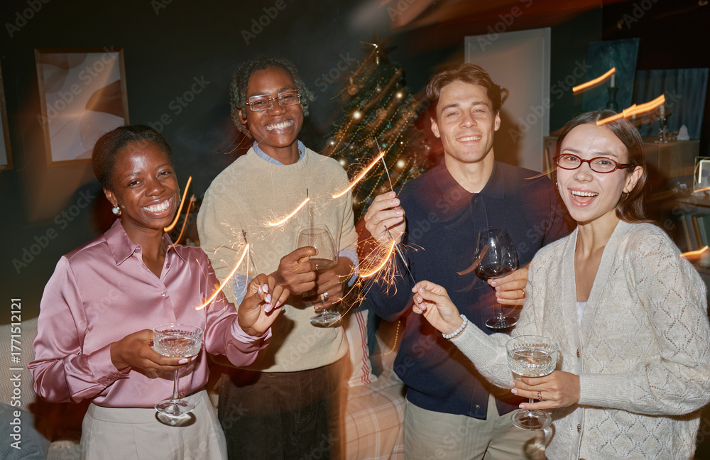 Obraz premium Group of young adult multiethnic friends celebrating together holding sparklers and drinks smiling at camera, standing indoors with Christmas tree in background during festive gathering