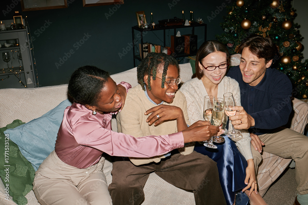 Obraz premium Diverse group of young adults sitting on sofa clinking champagne glasses, celebrating together near decorated Christmas tree, smiling and enjoying festive moment indoors