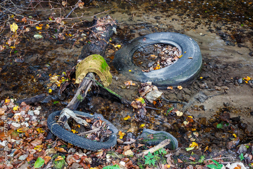 An unsightly mess of discarded tires, a boot, and other rubbish litters a shallow stream, highlighting environmental pollution amid autumn leaves