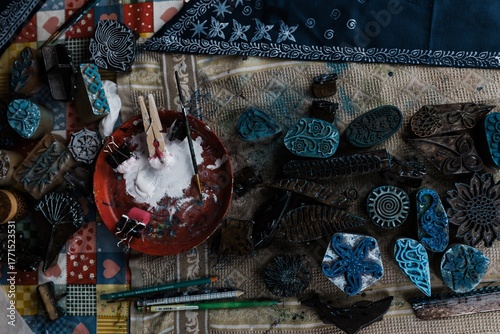 Fabric printing tools are laid out on the table: wooden carving stamps/blocks with patterns (block printing), brushes, a bowl of white paint/starch, clothespins and several pencils