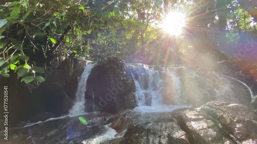Water rushes over rocks as bright sunlight creates visible flares and tiny droplets sparkle in the air, blending motion and light in a lush tropical scene. Waterfall light, tropical atmosphere.