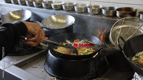 Chef stir frying Asian noodles with vegetables in hot wok at restaurant kitchen, with steam and flame.