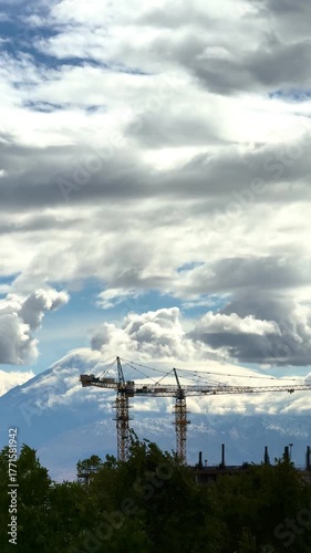 Two high-rise construction cranes on the construction of two skyscrapers against the backdrop of Mount Ararat and clouds during the day. The concept of modern construction processes and construction t