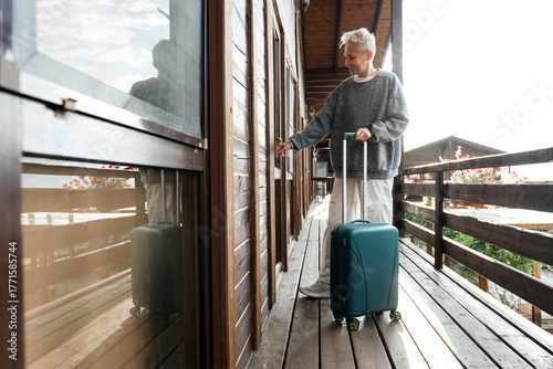 A young, stylish woman with a suitcase opens the door to a house. It's a trip or a business trip. Wooden floors and handrails.