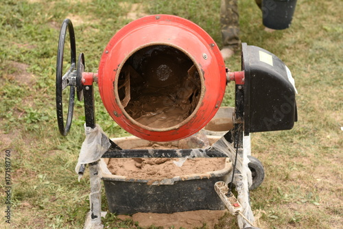 Wallpaper Mural Red cement mixer standing on grass, containing brown clay mixture inside. A container with mixed clay is visible underneath, showing the process of mixing clay for construction. Outdoor daylight setti Torontodigital.ca