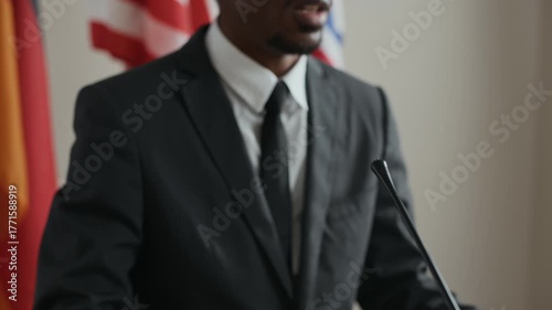 Tilt up of confident Black male politician in suit speaking at podium with microphones, addressing press and audience during formal political or international event