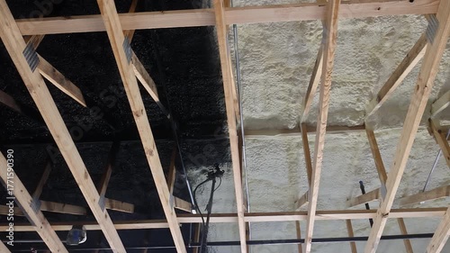 Worker on ladder is spraying black paint over foam insulation on wooden beams during renovation.