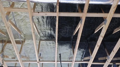 Contractor in protective gear applies refractory paint insulation using spray gun to wooden ceiling beams at construction site.
