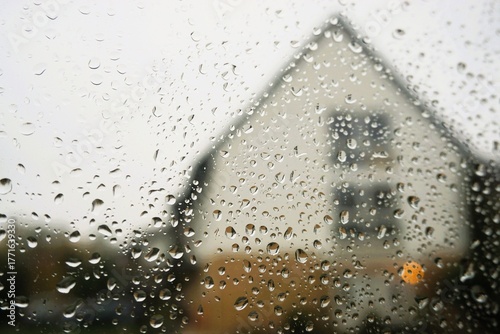Abstraktes Motiv mit Regentropfenmuster auf Glasscheibe vor Einfamilienhaus mit Spitzdach und weißem Himmel mit Licht bei Regen und Sturm am Morgen im Herbst