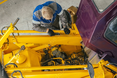 A mechanic in work gloves examines the engine of heavy machinery in a workshop. The setting is industrial, showcasing maintenance and repair work.