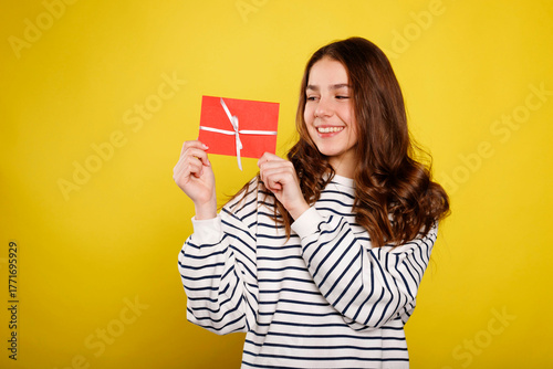 Young woman with long brown hair, wearing a striped sweater, joyfully holds a red gift envelope against a bright yellow background, celebrating a special occasion with excitement and happiness