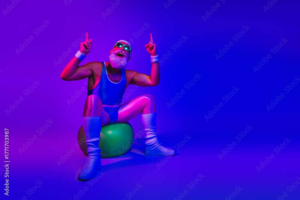 Fototapeta premium Retro fitness man in bright blue jumpsuit sits on a green exercise ball under neon purple and blue lights posing with raised fingers