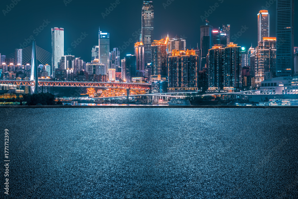 Fototapeta premium Empty asphalt road pavement with modern city skyline and illuminated commercial buildings at night.