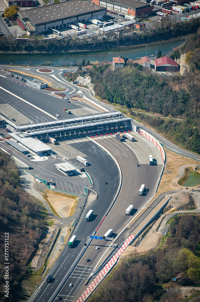 Fototapeta premium Aerial view of Biriatou showcasing a highway winding through green hills alongside the Bidassoa River. The scene includes industrial areas, residential buildings, and picturesque landscapes.