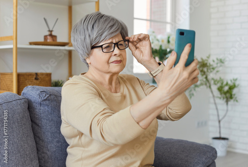 Portrait of a elderly senior woman sitting on sofa at home holding eyeglasses and trying to read message on smartphone having farsightedness eye disease. Presbyopia and vision problems concept.